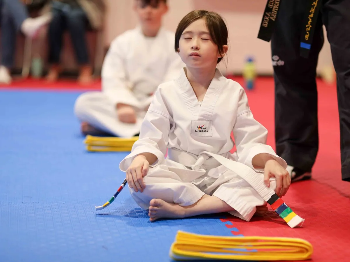 White belt student sitting cross-legged with her eyes closed, her new yellow belt folded on the mat in front of her, at a belt test