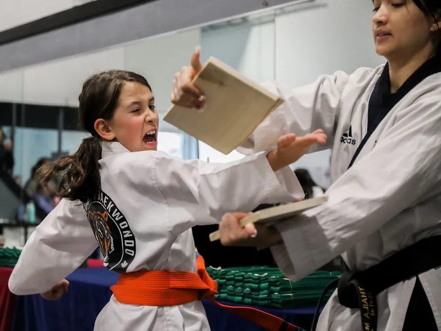 Orange belt student breaking a board with a palm strike and a full shout, her instructor holding the board, at a belt test