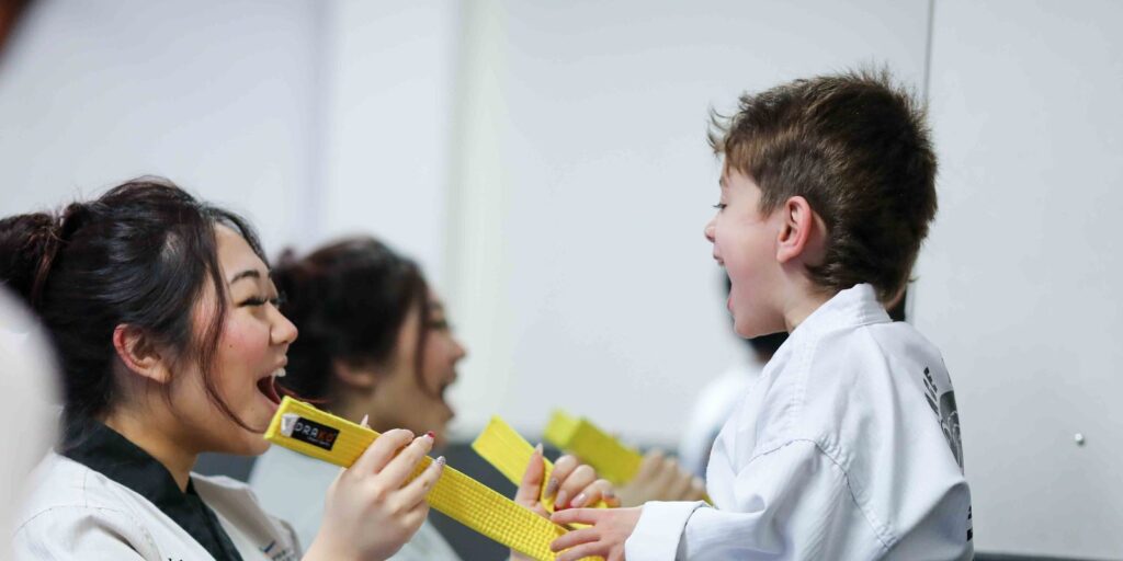A black belt helper laughs with a young yellow belt student over his belt at taekwondo camp