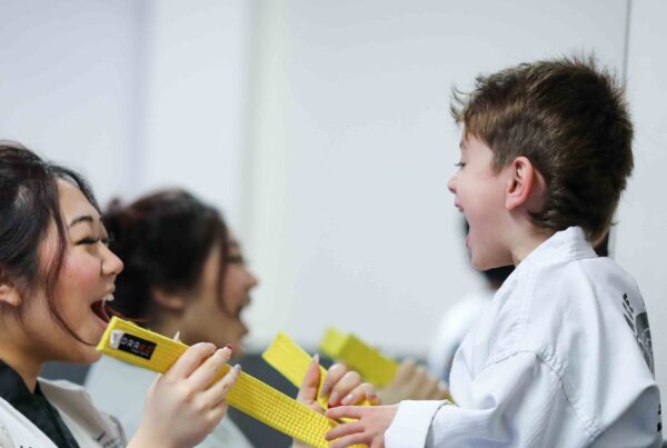 A black belt helper laughs with a young yellow belt student over his belt at taekwondo camp