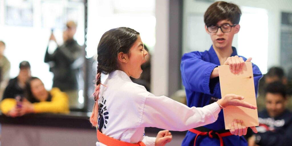 An orange belt student shouts as she breaks a wooden board with a palm strike at her belt test