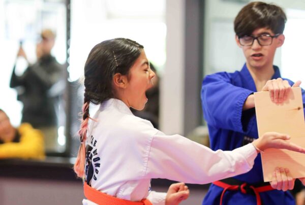 An orange belt student shouts as she breaks a wooden board with a palm strike at her belt test