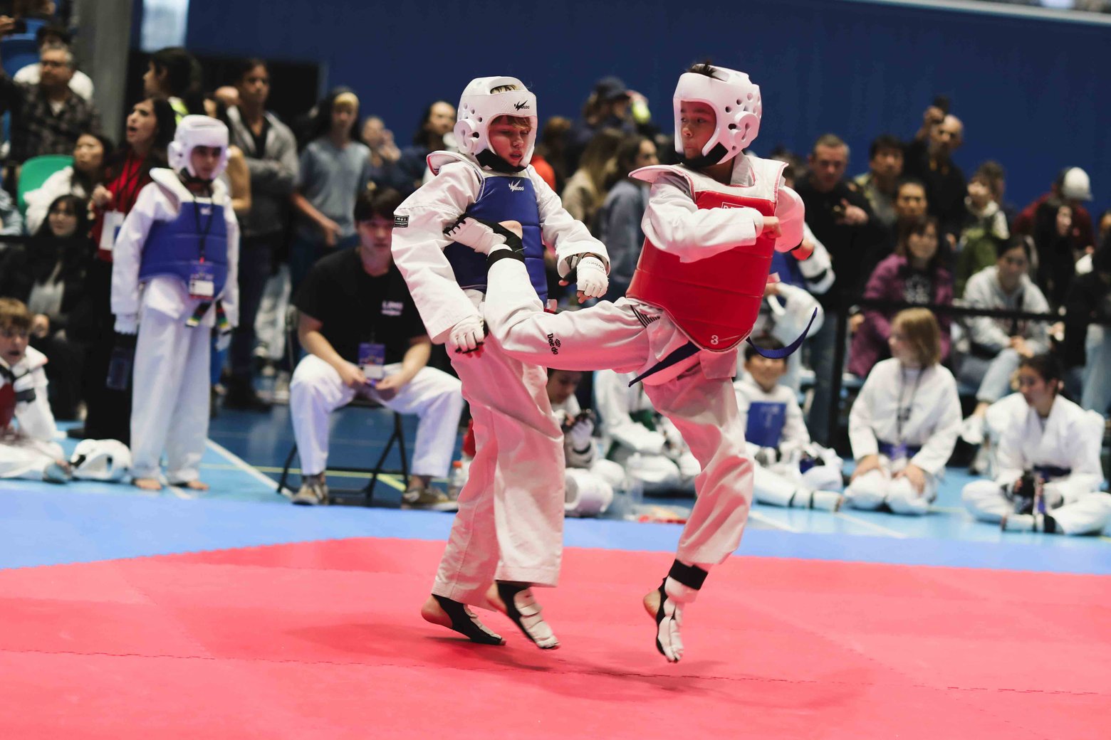 Two young Taekwondo students in full sparring gear, one landing a push kick on the other's chest protector at a tournament